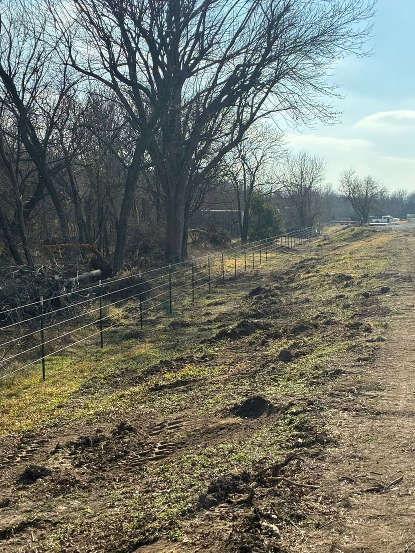 Rural landscape with a fence line, trees, and freshly disturbed soil under a blue sky.